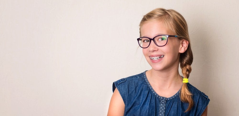 A young girl with glasses and braces, wearing a blue denim top, stands against a plain light-colored wall and smiles at the camera, showcasing the results of expert child orthodontics.