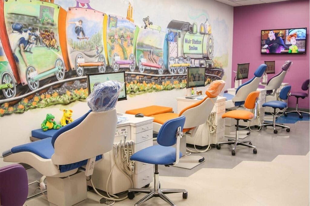 A children's dental clinic with colorful chairs, dental equipment, a wall mural of a train, and toys on the counter. A TV on the wall displays a cartoon.