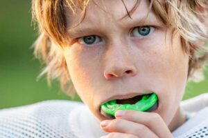 Close-up of a boy with light hair putting in a green mouthguard, wearing a white sports jersey or protective gear. Mouthguards help protect young athletes during intense play. Child wearing a sports mouthguard to help protect their teeth during sports.